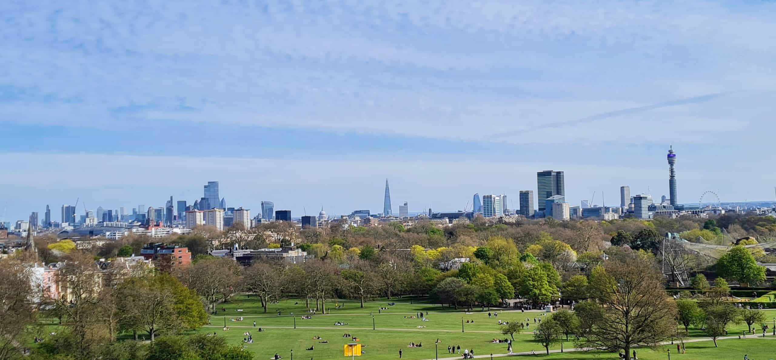looking at the skyline of london with all the tall buildings. in the forground is a park with lots of people on the grass.