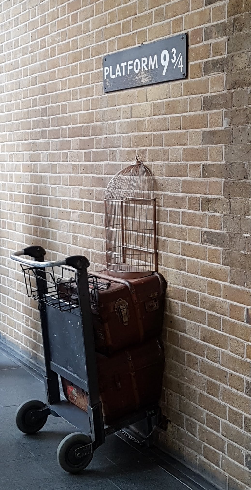 a picture of the Platform 3 3/4 sign from harry potter and a trolley with half a owl cage, and half a trunk going through a brick wall.