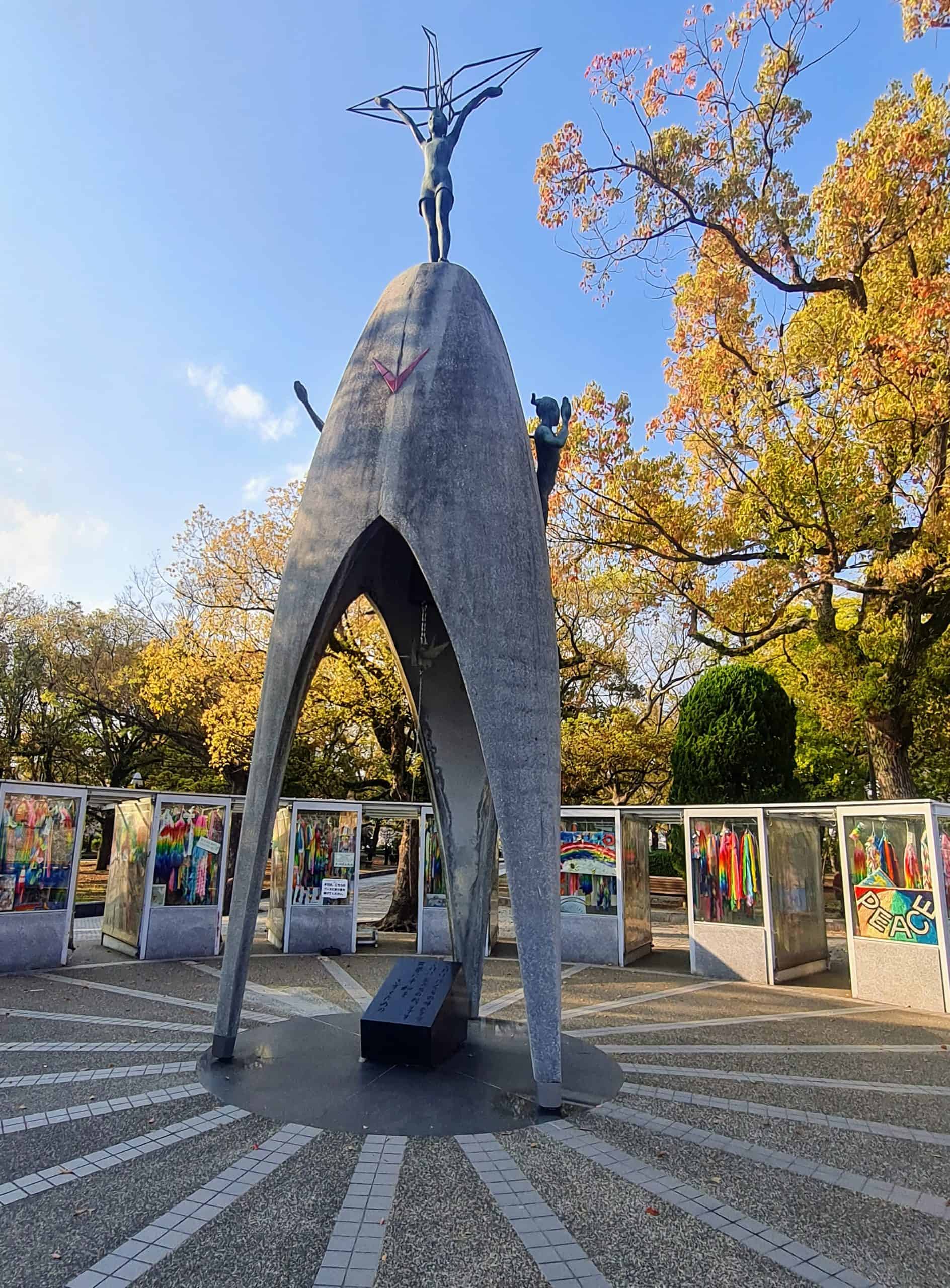 A large concrete dome shape with a statue of Sadako on the top with her holding her arms up, behind her is the shape of a paper crane