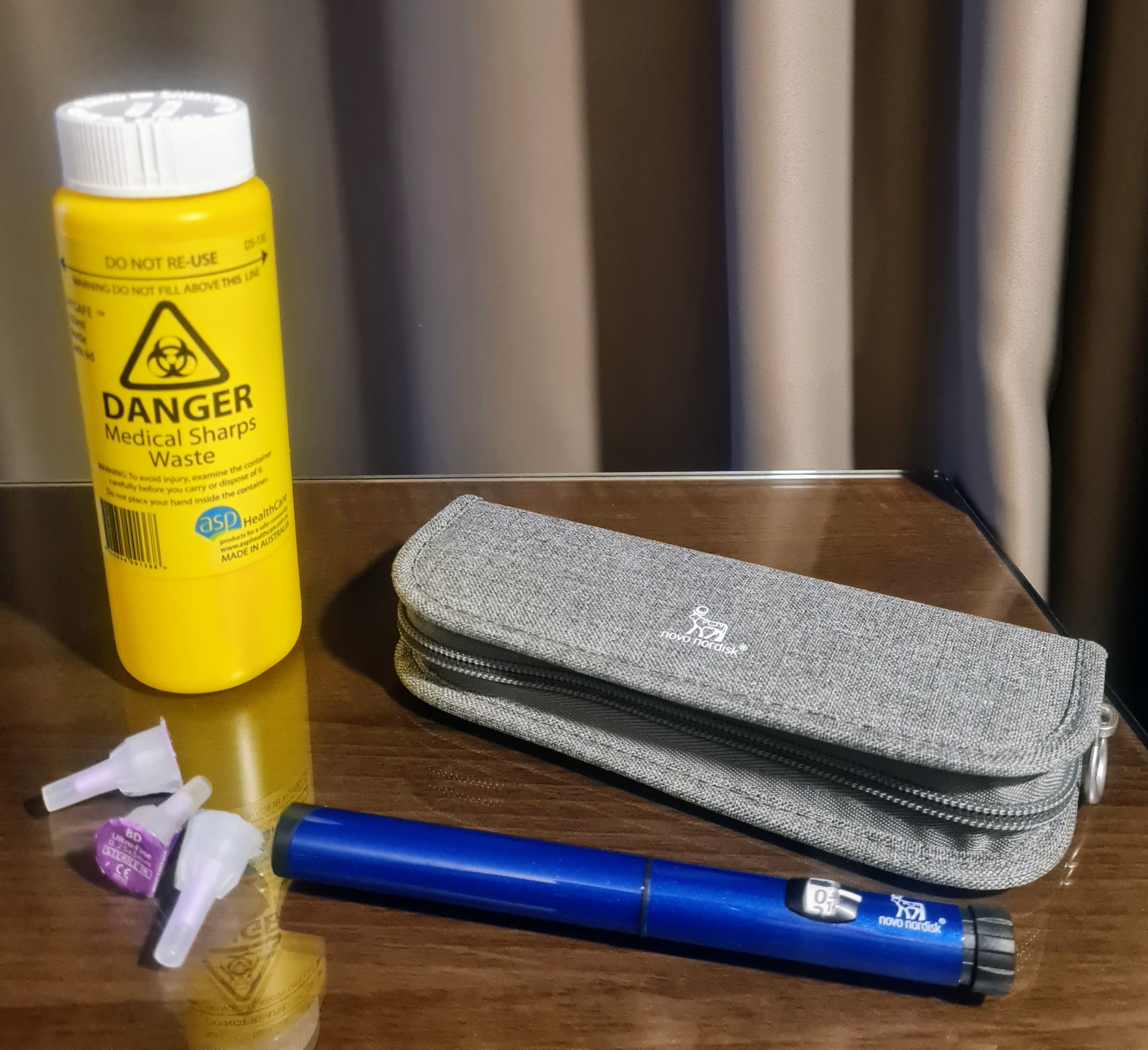 a collection of insulin essentials with a yellow sharps canister in the background, a small collection of needles, a grey zippered pouch and a insulin pen