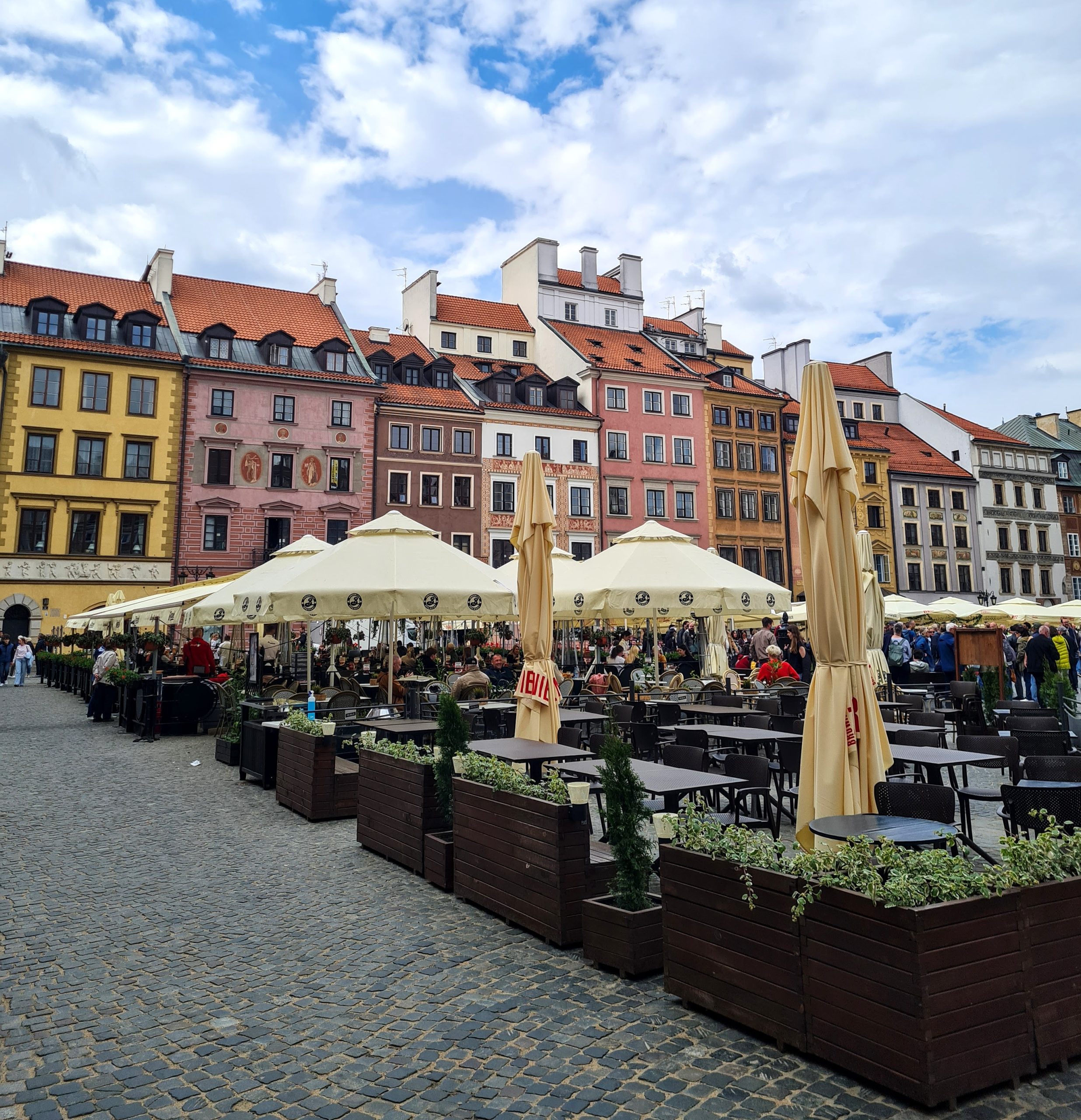 a photo of the Warsaw old town market square with colourful building facades in the background and umbrella's and seating for all the restaruants in the market square