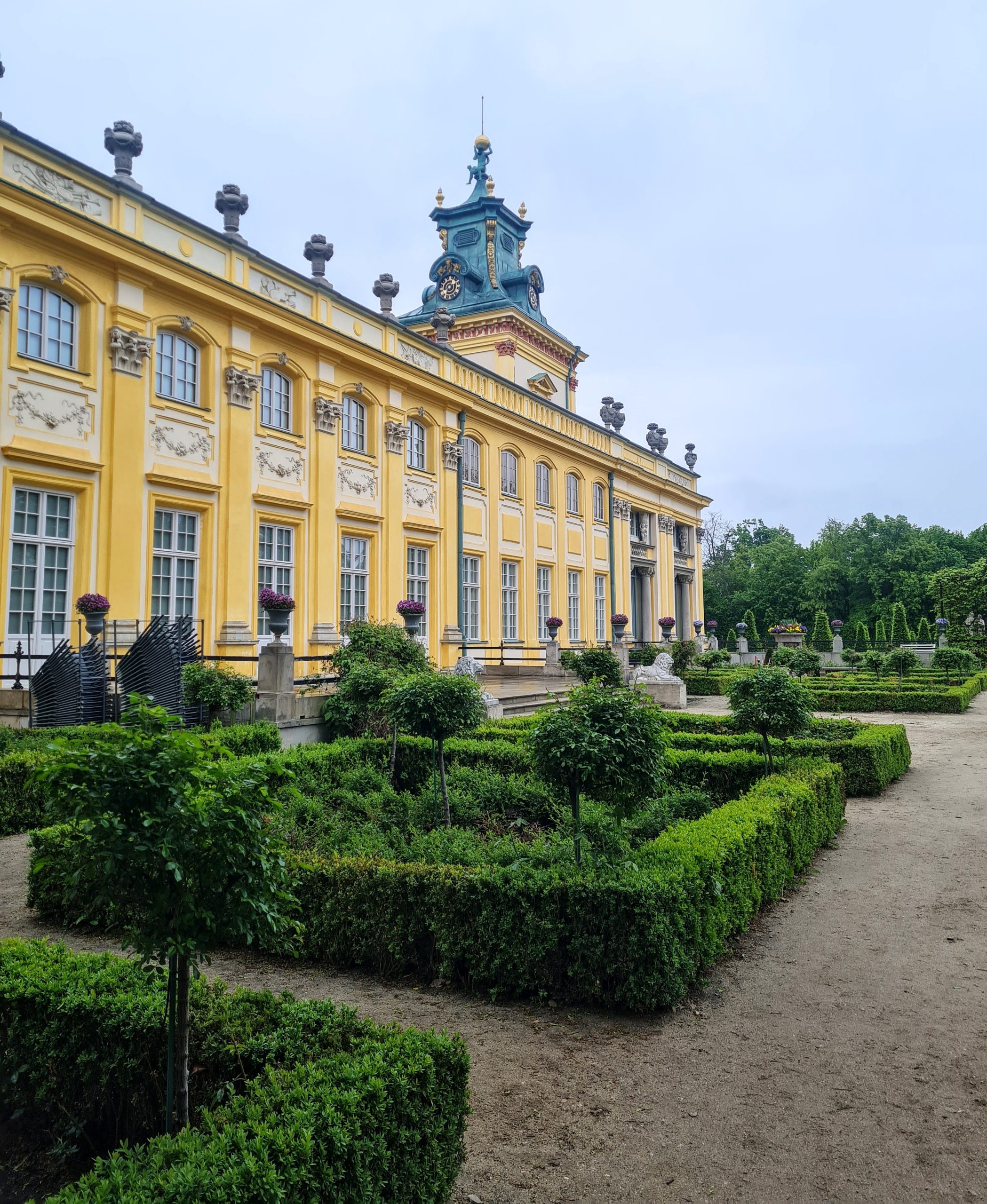 looking at the back of Wilanow palace with the green hedges in the foreground and the pastal yellow facade of the palace dotted with windows in the background