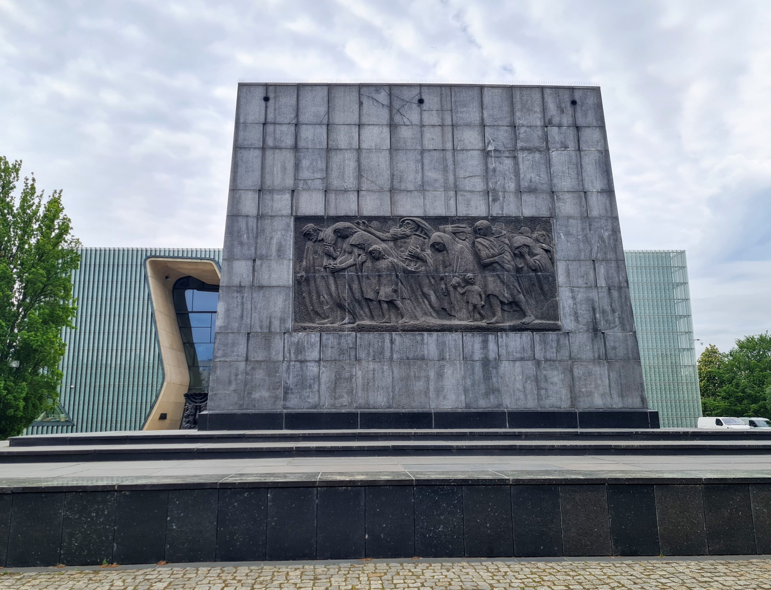 a photo of the Warsaw ghetto memorial with a carving of men, women and children all in a line into a large grey block of granite about 20 metres tall